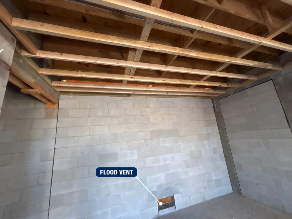 Ground level enclosure beneath an elevated coastal home in a Florida V zone, showing the elevated floor framing above, breakaway wall construction, and a labelled flood vent opening at the base of the wall