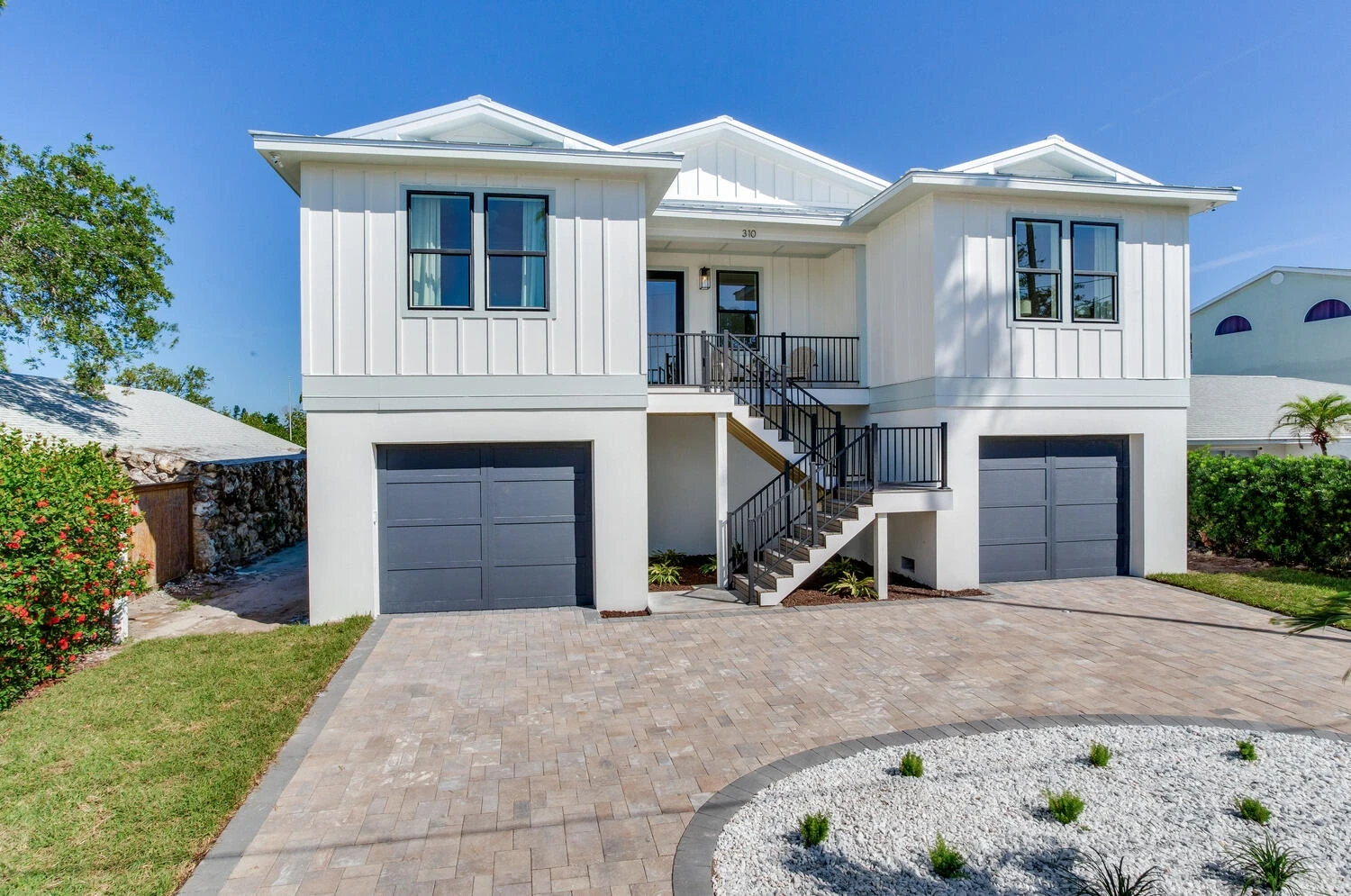 Front elevation of a two-story elevated coastal home with an exterior stair and two garage doors above a paver driveway